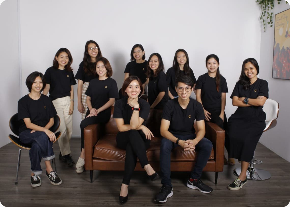 Twelve coworkers in black shirts posing together in a bright, modern office space around a leather sofa.